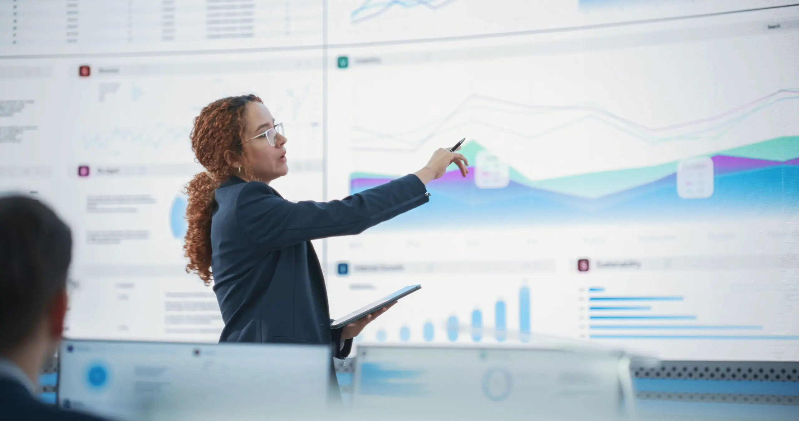 Female Senior Data Scientist Reviewing Reports On Big Digital Screen In Monitoring Room. Diverse Consulting Company Employees Working Behind Desktop Computers.